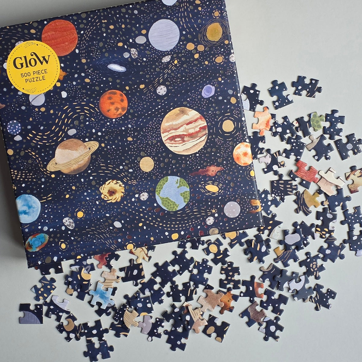 A jigsaw puzzle box decorated with images of planets and stars, with scattered jigsaw pieces on a grey background.