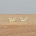 A pair of small gold butterfly stud earrings displayed on a light wood surface against a soft gray background.