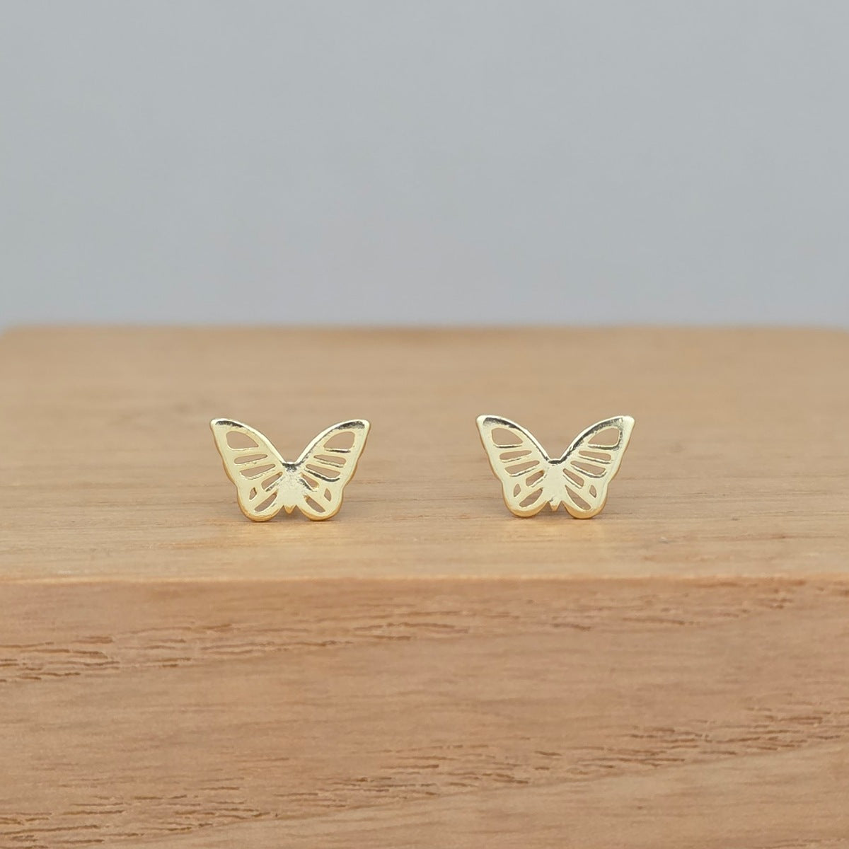 A pair of small gold butterfly stud earrings displayed on a light wood surface against a soft gray background.