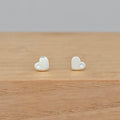 A pair of small heart-shaped stud earrings in a pale gold or cream tone, each with a tiny clear stone, displayed on a light wood surface against a soft gray background.