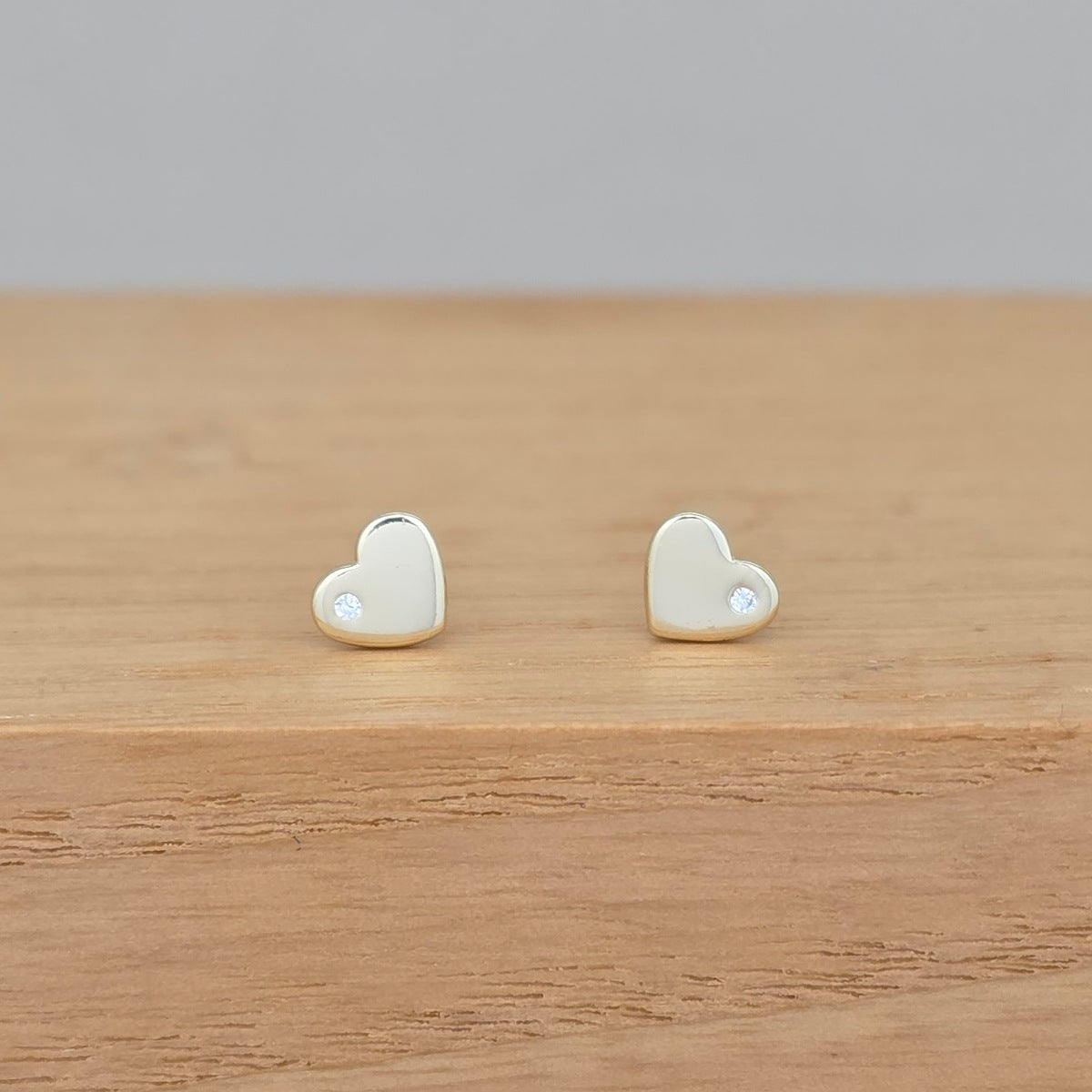 A pair of small heart-shaped stud earrings in a pale gold or cream tone, each with a tiny clear stone, displayed on a light wood surface against a soft gray background.
