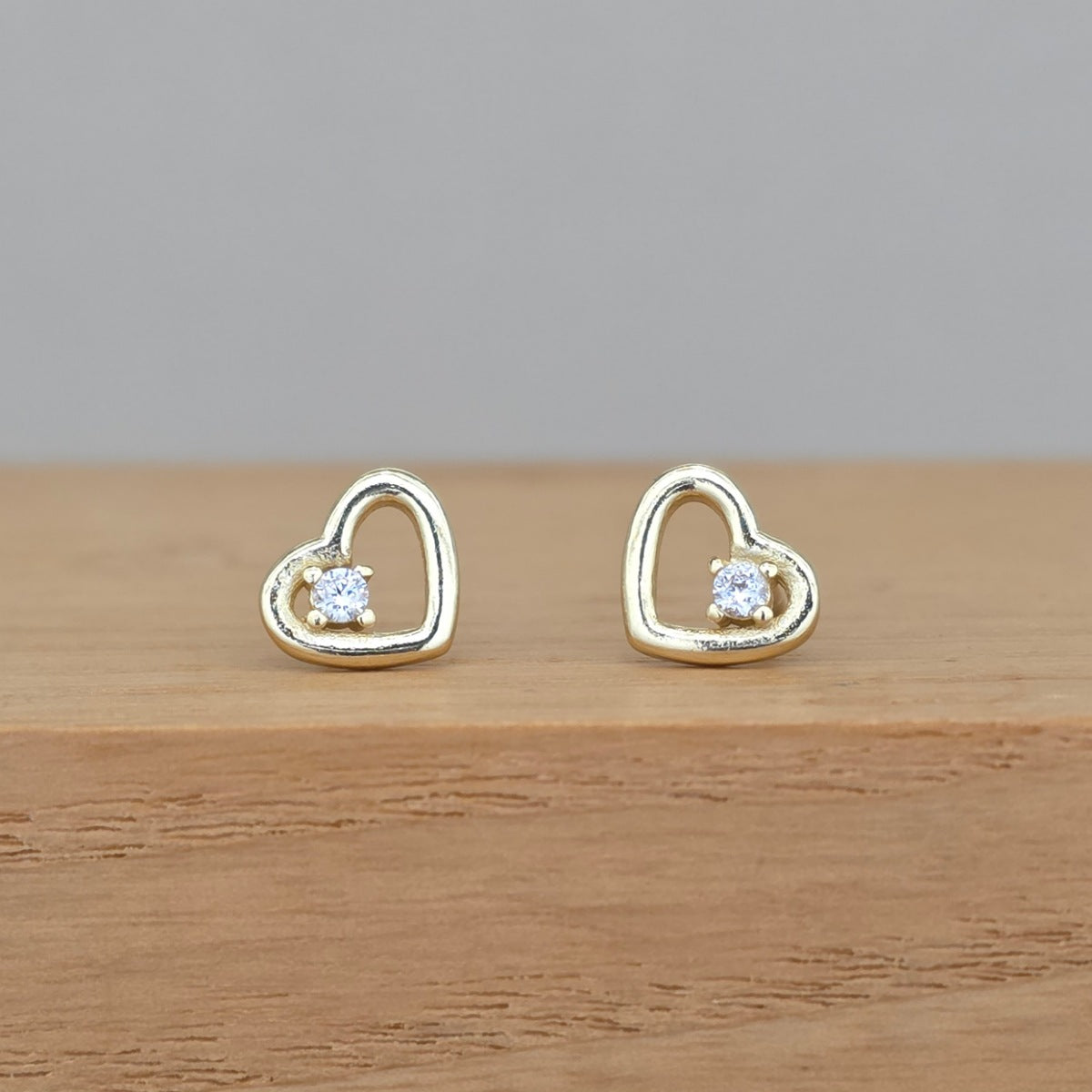 A pair of gold heart-shaped stud earrings with a small clear gemstone in the center, displayed on a wooden surface against a neutral gray background.