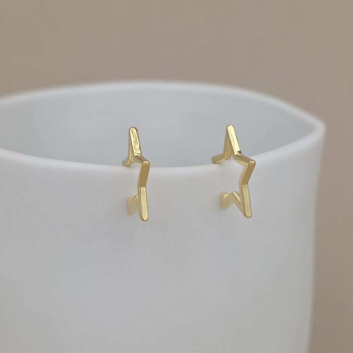 A pair of gold star hoop earrings, displayed on the rim of a white ceramic cup against a soft beige background.