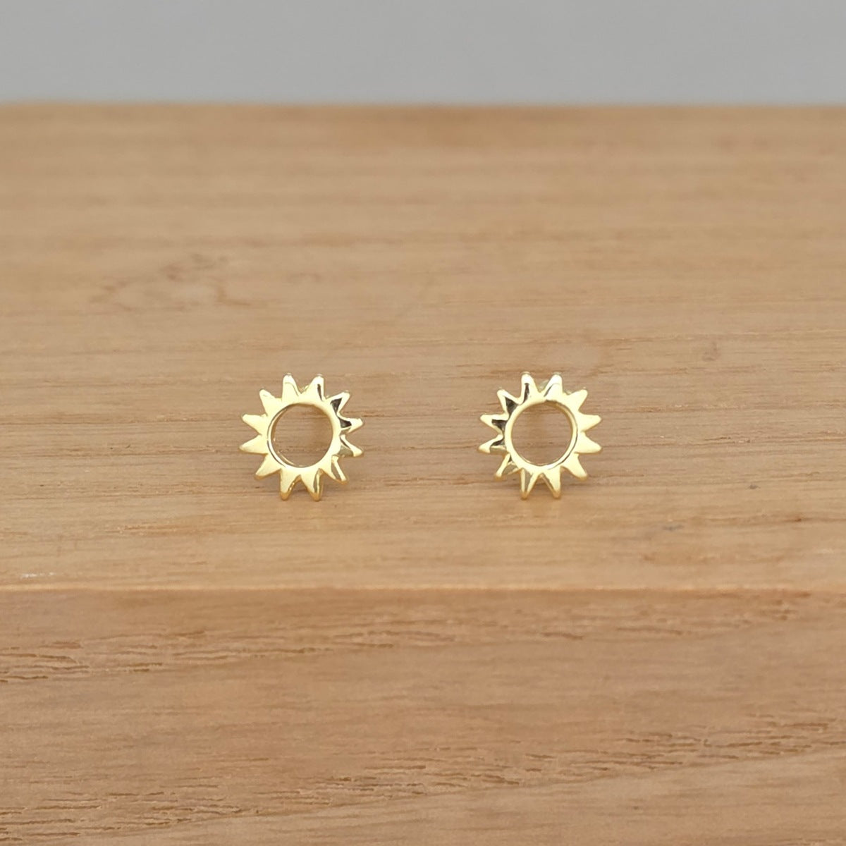 A pair of gold sunburst stud earrings resting on a light brown wooden surface.