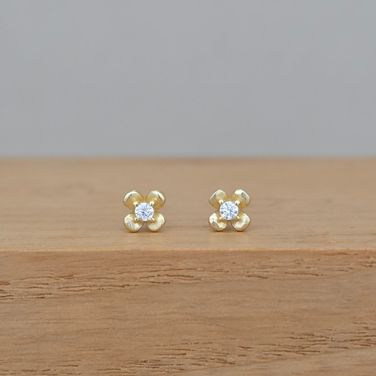A pair of small gold flower stud earrings with clear central gemstones, resting on a light wood surface against a soft gray background.