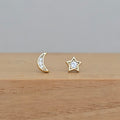 A pair of gold-toned stud earrings, one shaped like a crescent moon and the other like a five-pointed star, both set with small clear stones, resting on a light wooden surface against a neutral gray background.