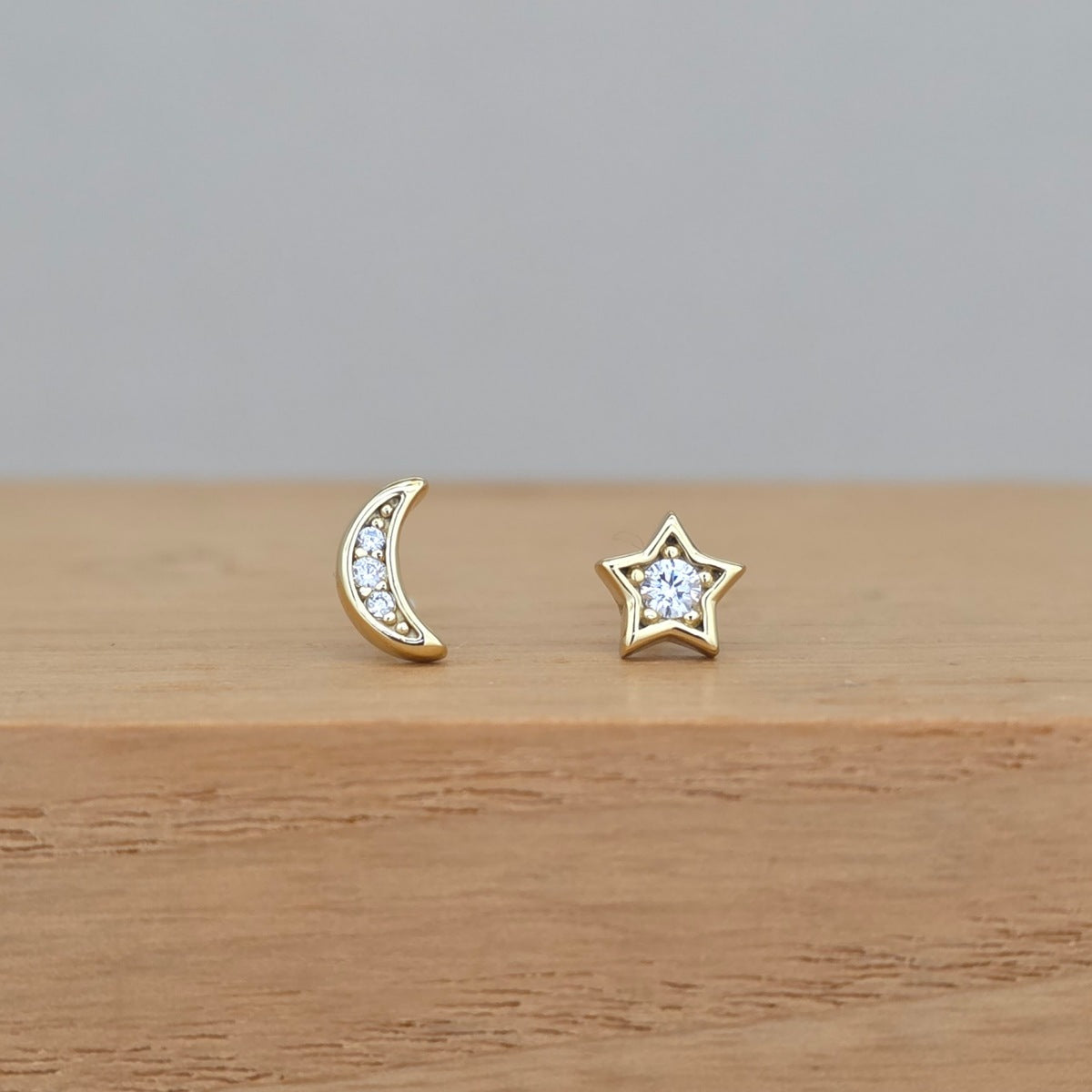 A pair of gold-toned stud earrings, one shaped like a crescent moon and the other like a five-pointed star, both set with small clear stones, resting on a light wooden surface against a neutral gray background.