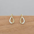 A pair of gold teardrop-shaped drop earrings with clear pavé stones along the lower curve, displayed on a wooden surface against a neutral gray background.