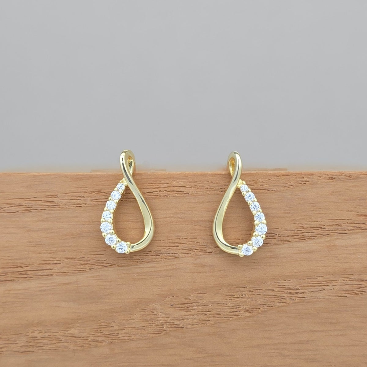 A pair of gold teardrop-shaped drop earrings with clear pavé stones along the lower curve, displayed on a wooden surface against a neutral gray background.
