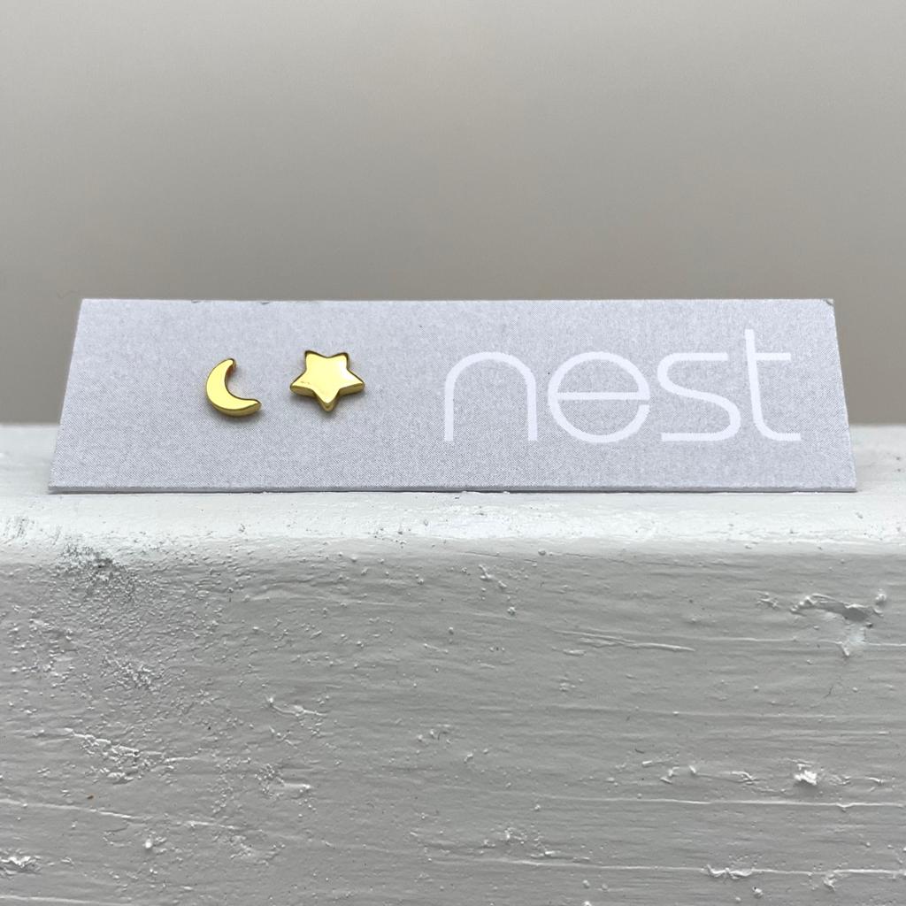 A pair of small gold crescent moon and star stud earrings displayed on a light gray branded card that reads 'nest', resting on a textured white surface against a soft gray background.