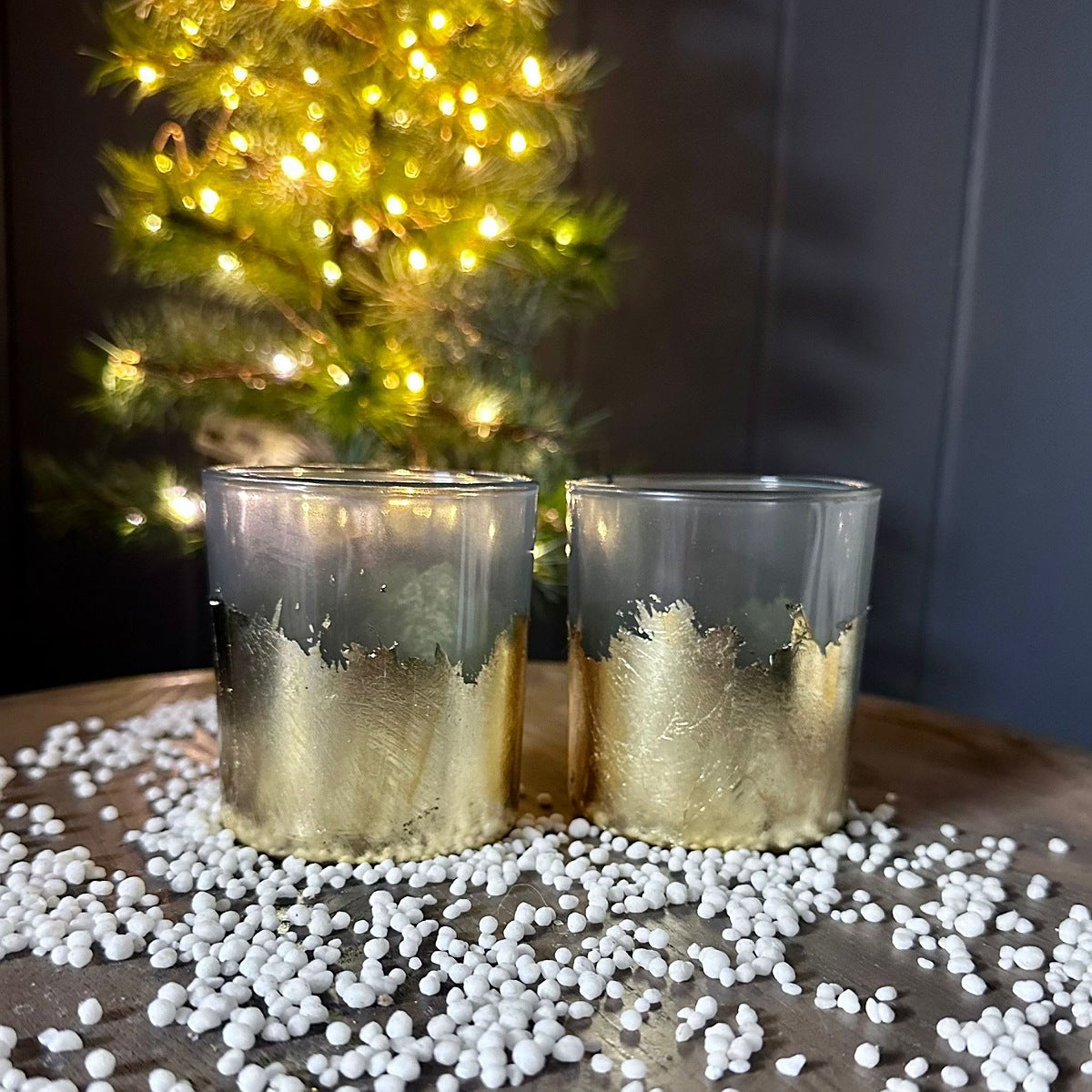 Two clear glass candle holders with a metallic gold leaf pattern at the bottom, styled on a wooden surface with white decorative beads and a blurred Christmas tree in the background.