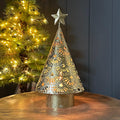 A decorative gold metal Christmas tree tea light holder with intricate laser-cut patterns on a galvanised base, lit with warm yellow light, set on a wooden table with a blurred lit Christmas tree in the background.