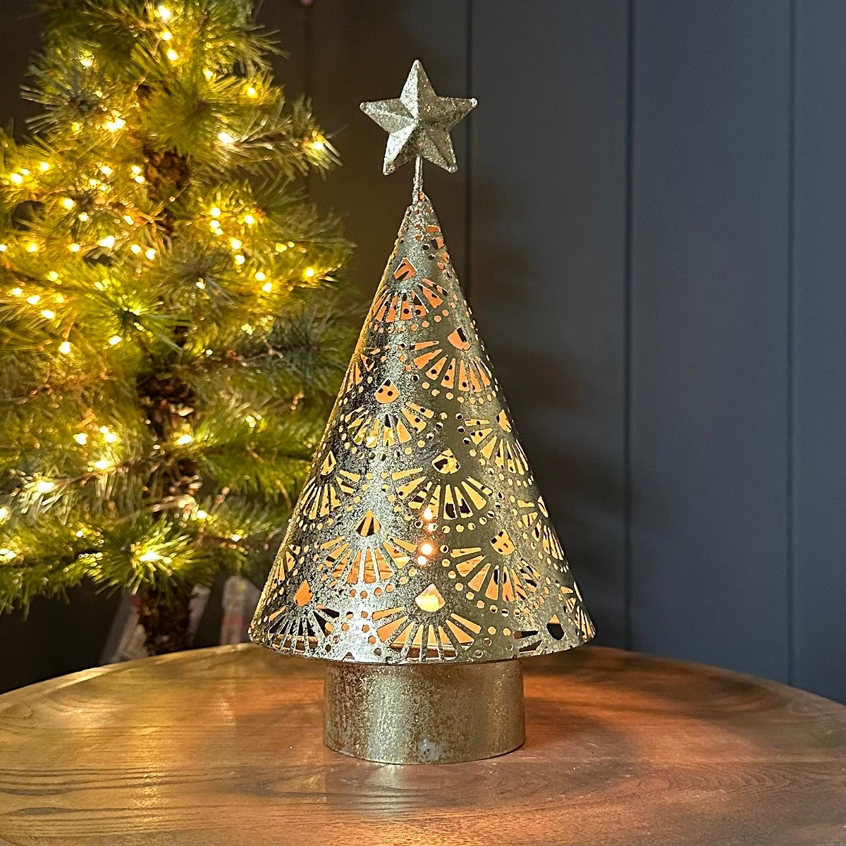 A decorative gold metal Christmas tree tea light holder with intricate laser-cut patterns on a galvanised base, lit with warm yellow light, set on a wooden table with a blurred lit Christmas tree in the background.