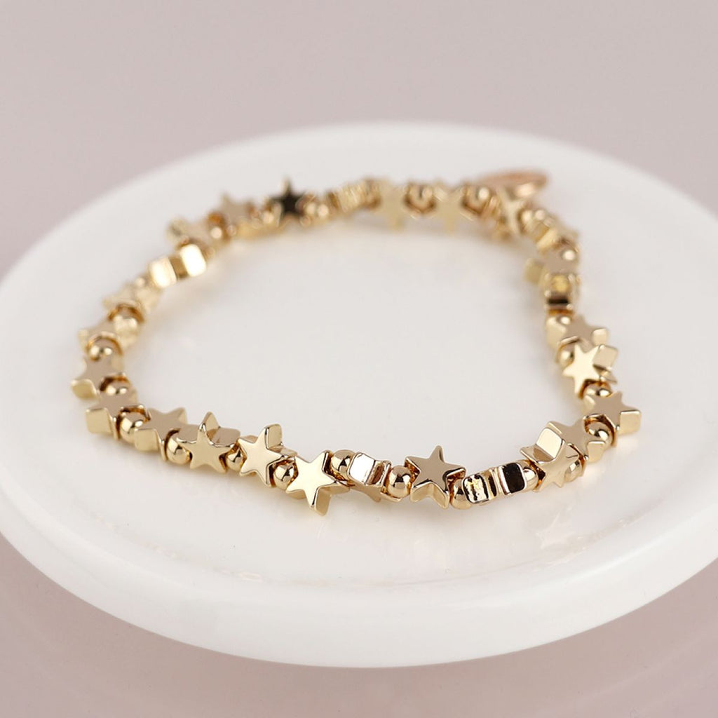A gold bracelet with connecting star and bead charms, resting on a white ceramic dish against a warm beige background.