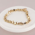 A gold bracelet with connecting star and bead charms, resting on a white ceramic dish against a warm beige background.