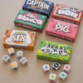 Collection of board game boxes including 'Eggsactly Six', 'Greedy Pig', 'Dicey Words', and others, with dice on a beige surface.