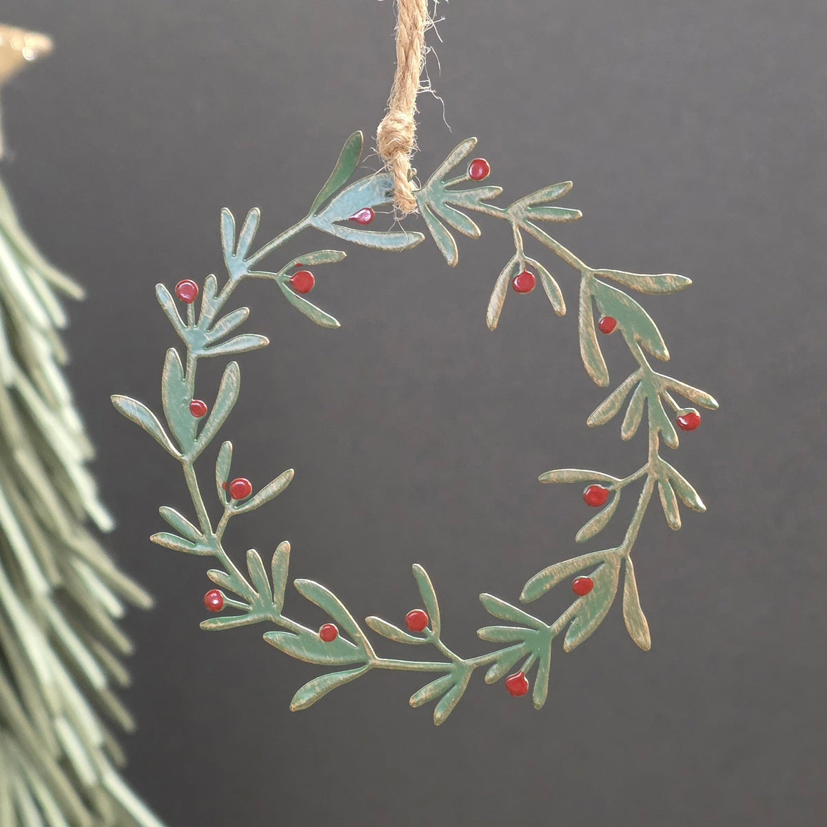 A metal wreath Christmas ornament with a distressed green and gold finish, decorated with small red berries, hanging by a jute twine string against a dark grey background.
