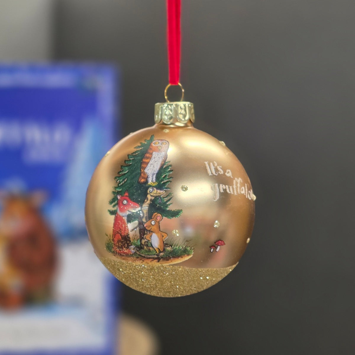 A round gold Christmas ornament with a red hanging ribbon, featuring a festive illustration of woodland animals around a tree and the text 'it's a gruffalo'.