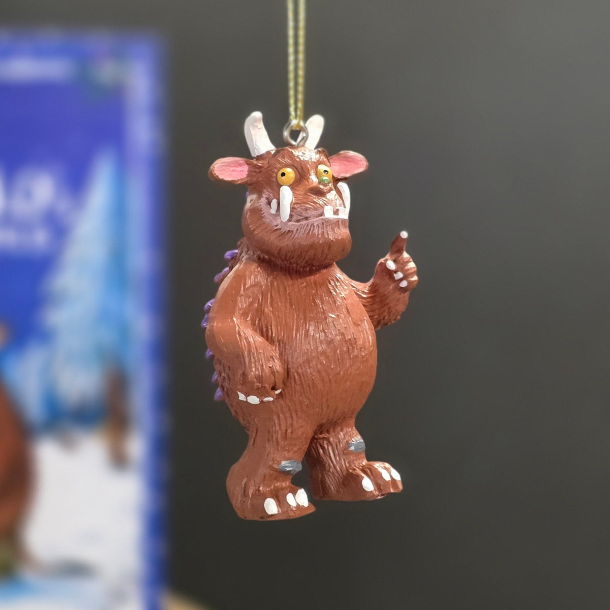 A brown Gruffalo Christmas ornament hanging from a gold string against a dark grey background with a blurred blue book in the side.