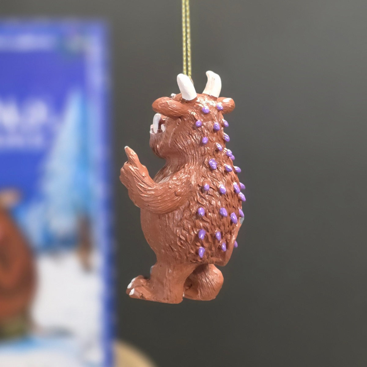 A small brown creature ornament with white horns and purple spines hanging by a gold string against a dark grey background with a blurred blue box behind it.