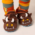 A pair of brown plush monster baby shoes with white horns, yellow eyes, and stitched teeth, shown on a child's feet with striped socks.