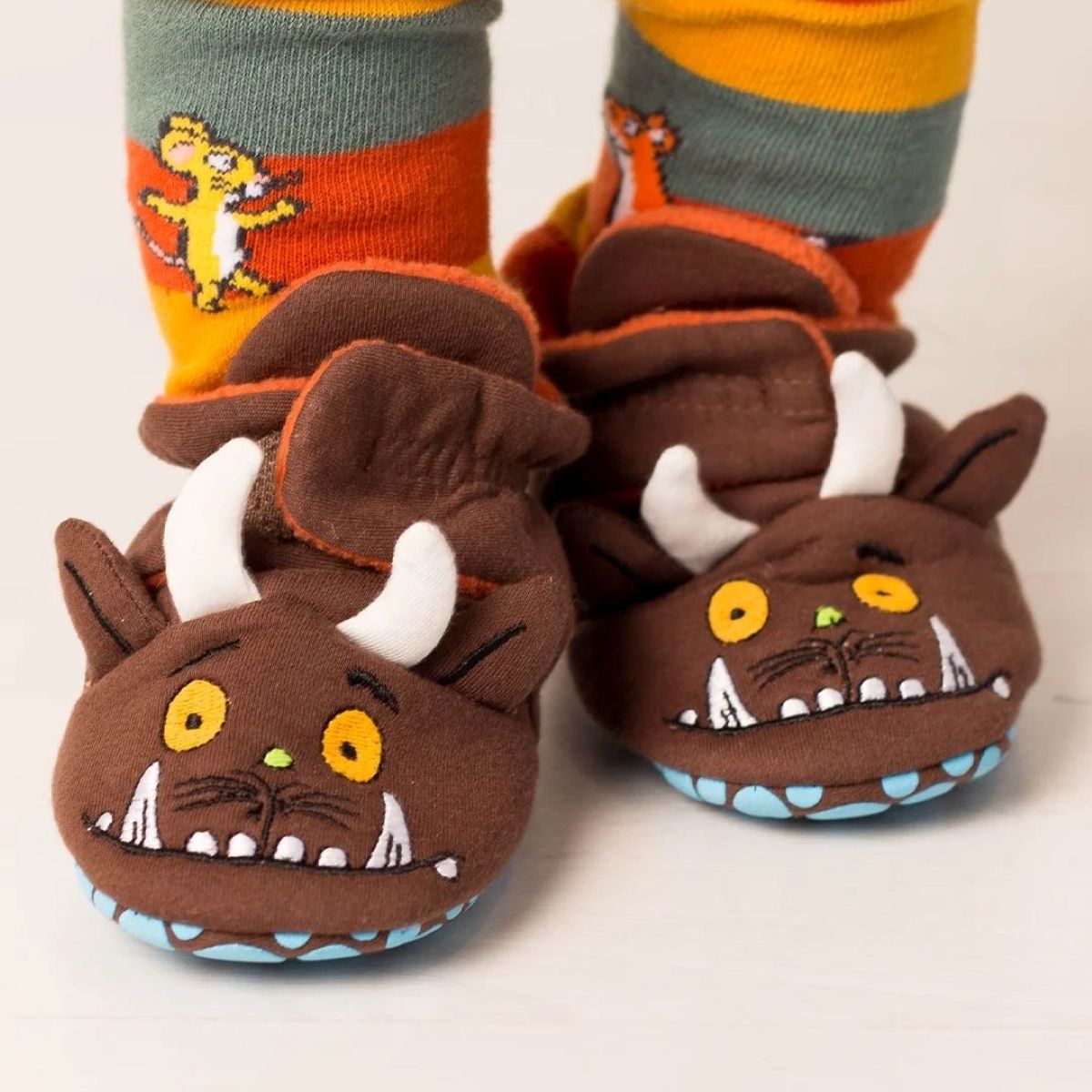 A pair of brown plush monster slippers with white horns, yellow eyes, and a blue stitched mouth, shown on a child's feet with striped socks featuring a cartoon character.