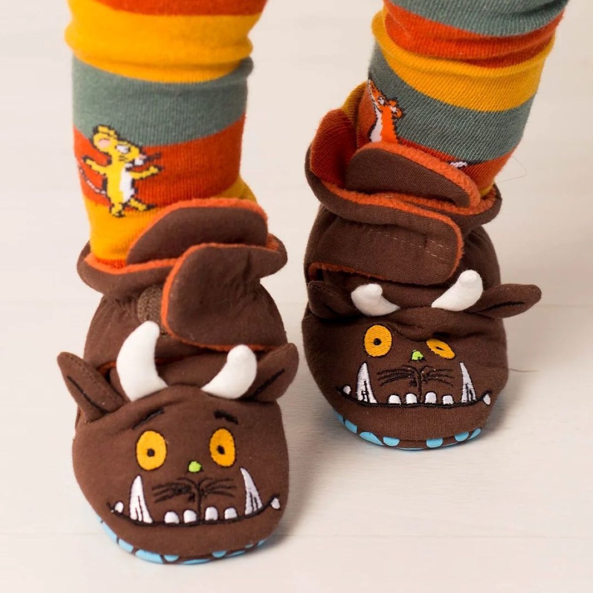 A close-up of a child's feet wearing brown monster-themed slipper shoes with white horns, yellow eyes, and blue soles, paired with colorful striped socks on a light wooden floor.