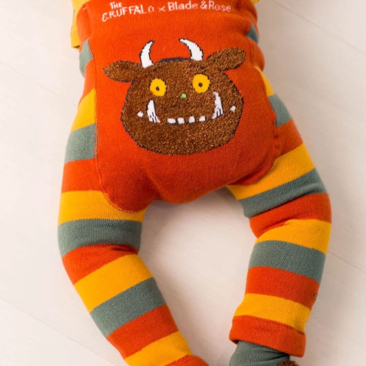 A baby's footed sleeper with orange and yellow stripes on the legs and a large brown monster face applique on the belly. The top is orange with white text reading 'The CRUFFALO x Blade & Rose'.