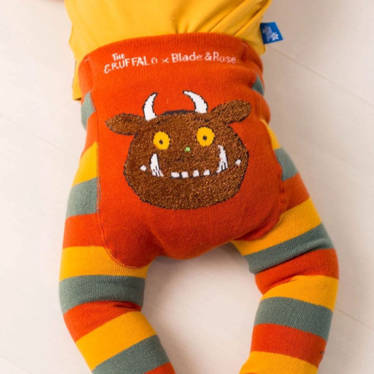 A close-up of a children's outfit featuring a bright orange waistband with embroidered text reading 'The Gruffalo x Blade & Rose' and a large brown monster face motif, paired with striped legs in orange, yellow, and gray-green on a white background.
