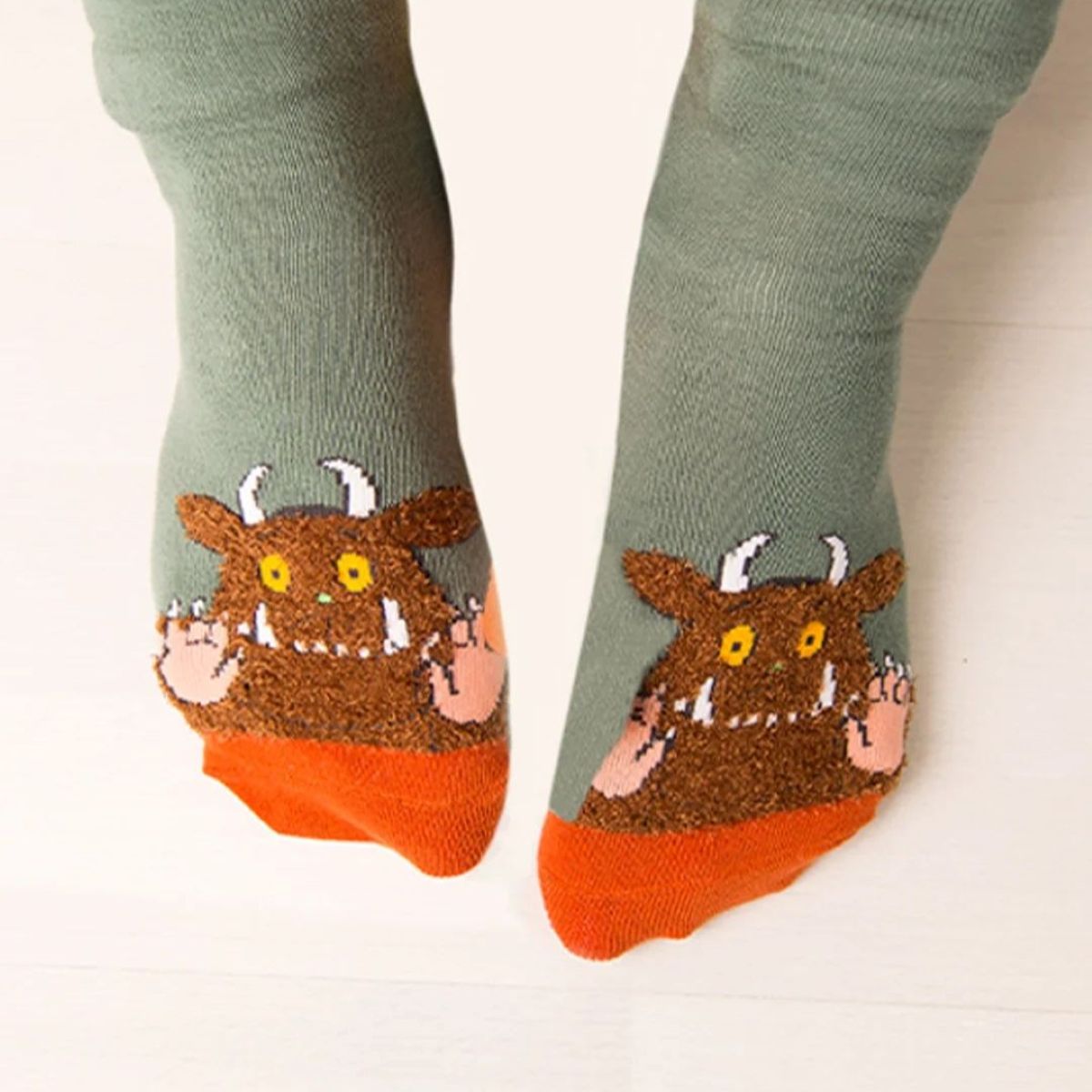 A pair of sage green socks with a brown monster face design on the foot and bright orange toes, shown worn on feet against a light wood floor.