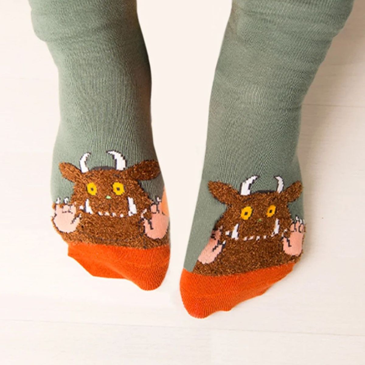 A pair of socks worn on feet, featuring a green upper leg section with a brown monster face graphic, an orange toe cap, and pink paw details.