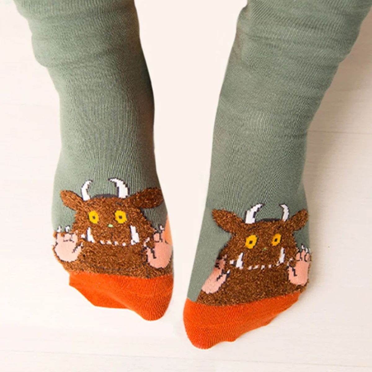 A pair of green socks worn on feet, featuring a brown monster design with yellow eyes, white horns, and pink claws on the foot, and an orange toe.