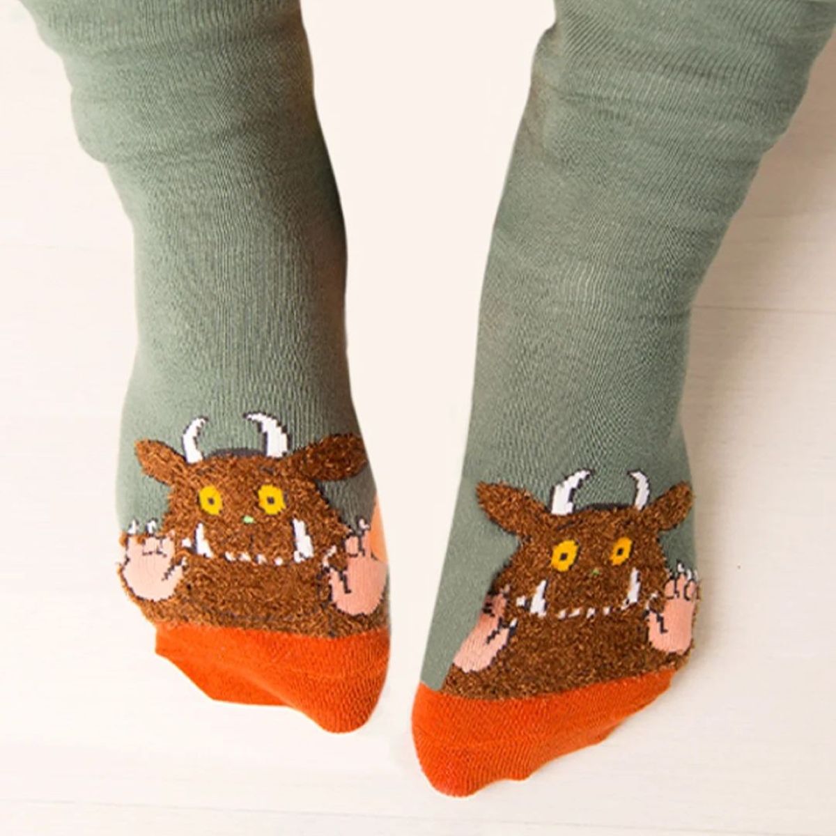 A pair of socks worn on feet, featuring a sage green upper, an orange toe cap, and a brown monster face with yellow eyes and white horns near the foot.