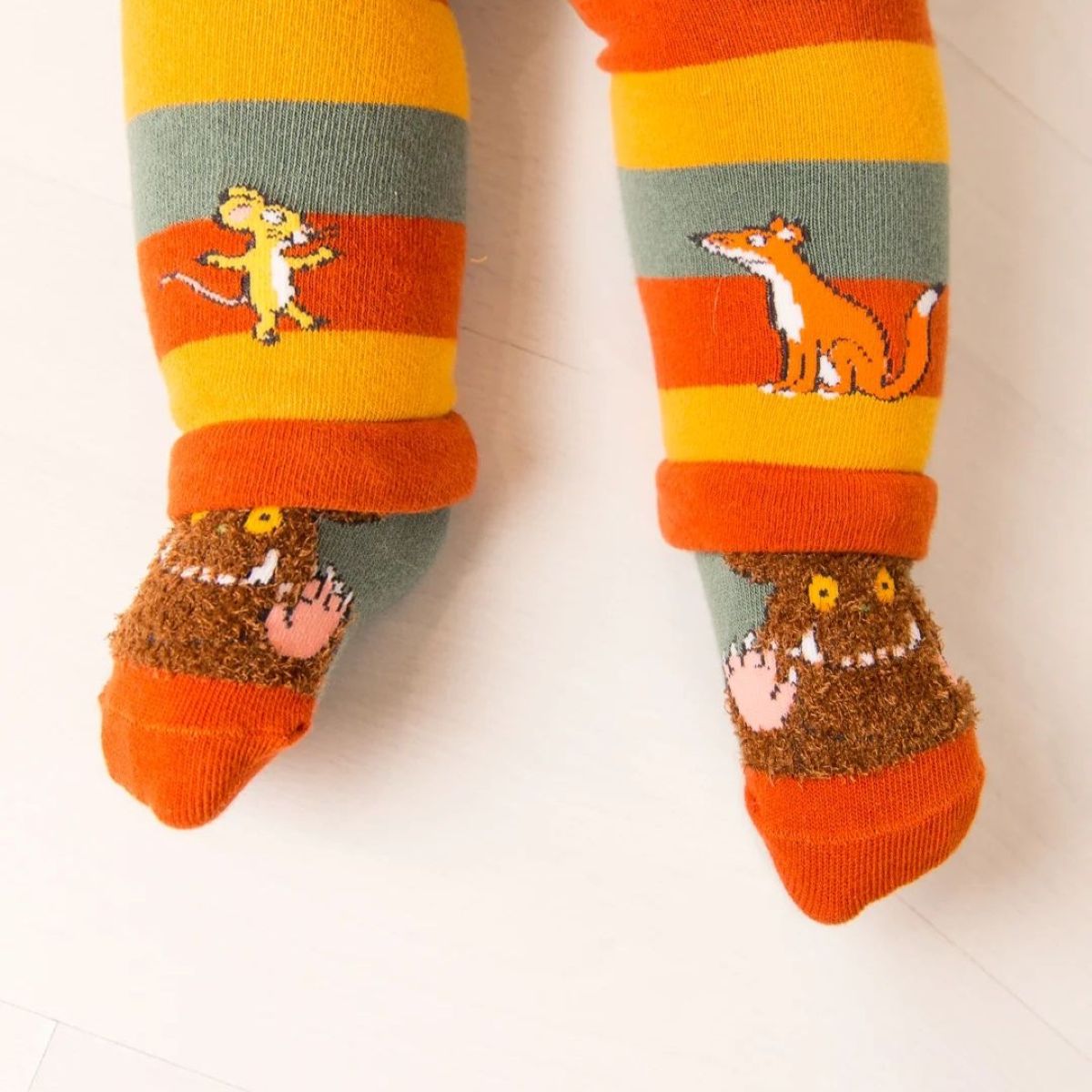 A pair of children's striped socks worn on a child's legs, featuring orange, yellow, and gray horizontal stripes with knitted animal characters and a brown fuzzy monster design on the feet.