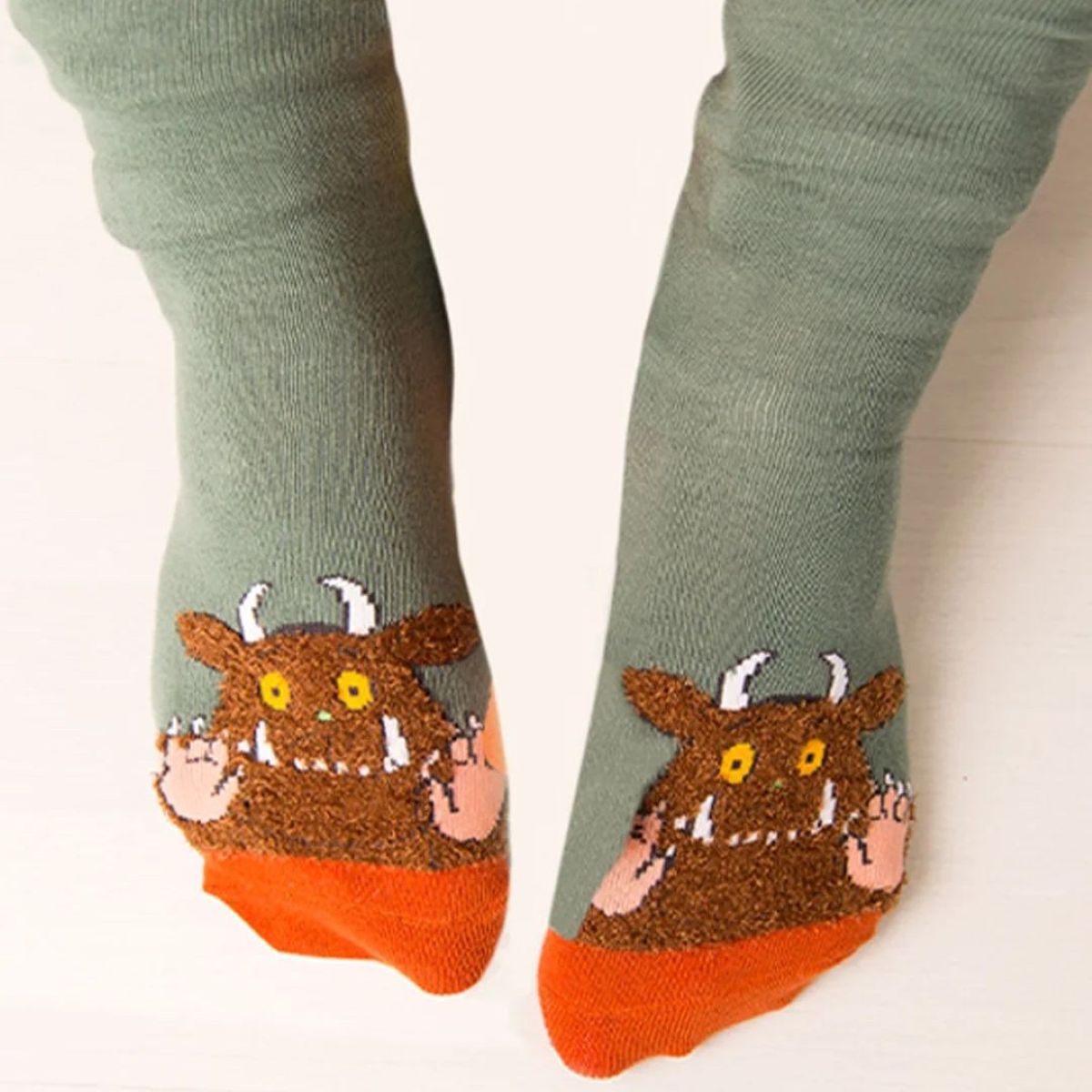 A pair of sage green crew socks with a brown monster graphic on the foot, featuring orange toes and yellow eyes, shown on feet against a light background.