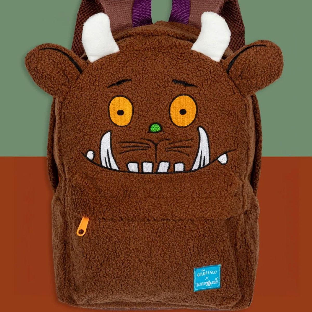 A brown plush monster backpack designed to look like a furry creature with white horns, yellow eyes, a green nose, and a wide white grin. The backpack has a front pocket with an orange zipper pull and a small blue 'Crazy Dog & Bunny' brand patch near the bottom right.