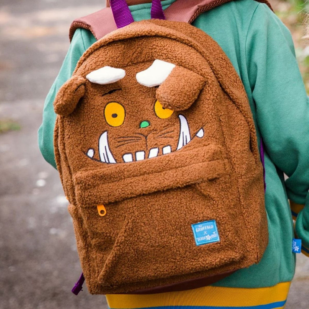 A brown plush monster face backpack worn by a person in a green sweatshirt, photographed outdoors on a paved path.