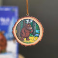 A circular wooden slice ornament featuring an illustration of the Gruffalo and mouse in the woods.  Hanging by a brown ribbon against a blurred background.
