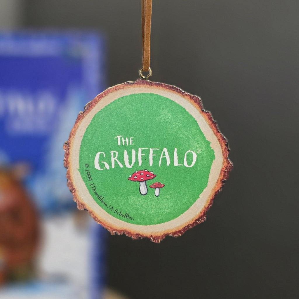 A round wooden slice hanging ornament featuring a green center with the text 'THE GRUFFALO' and small red-and-white mushrooms, suspended by a brown string against a blurred background.