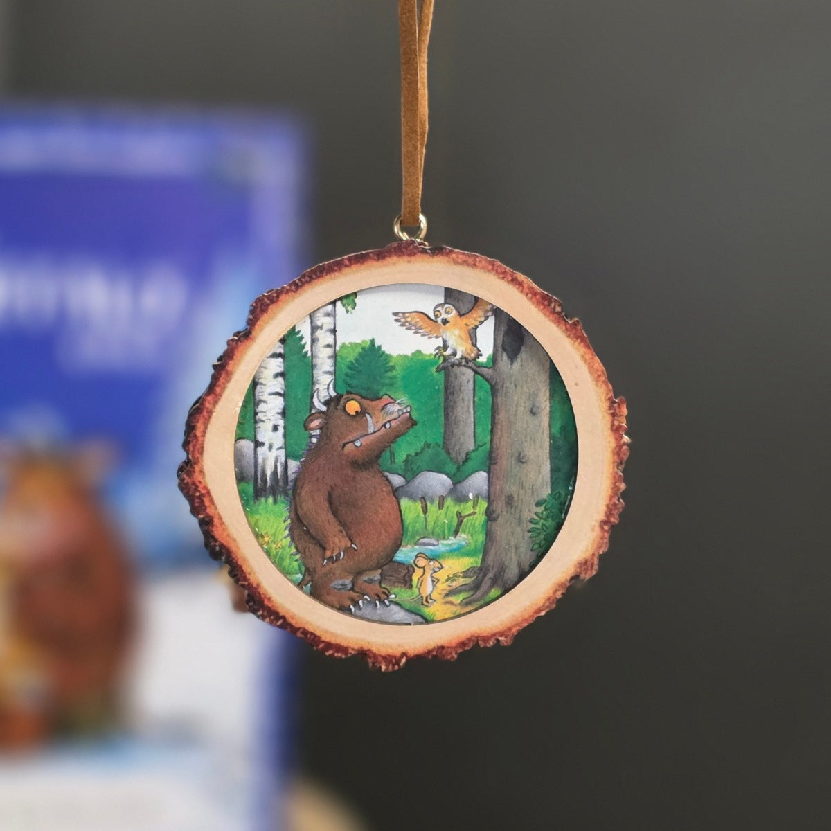 A round wooden slice ornament with a hanging loop, featuring an illustrated forest scene with the Gruffalo, birch trees, and an owl, photographed against a blurred dark background.