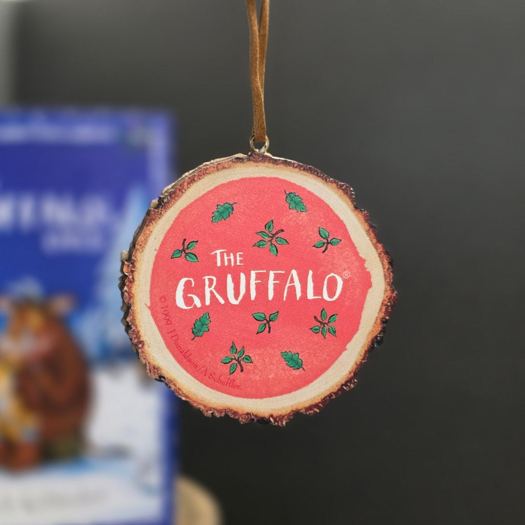 A round wooden slice ornament hanging by a brown string, featuring a red center with the white text 'The Gruffalo' and small green leaf illustrations, against a dark blurred background.