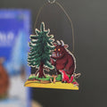 A handcrafted wooden hanging ornament featuring a whimsical illustration of the Gruffalo, a red fox, and a pine tree on a yellow forest floor, suspended by a thin metal wire against a dark grey background.