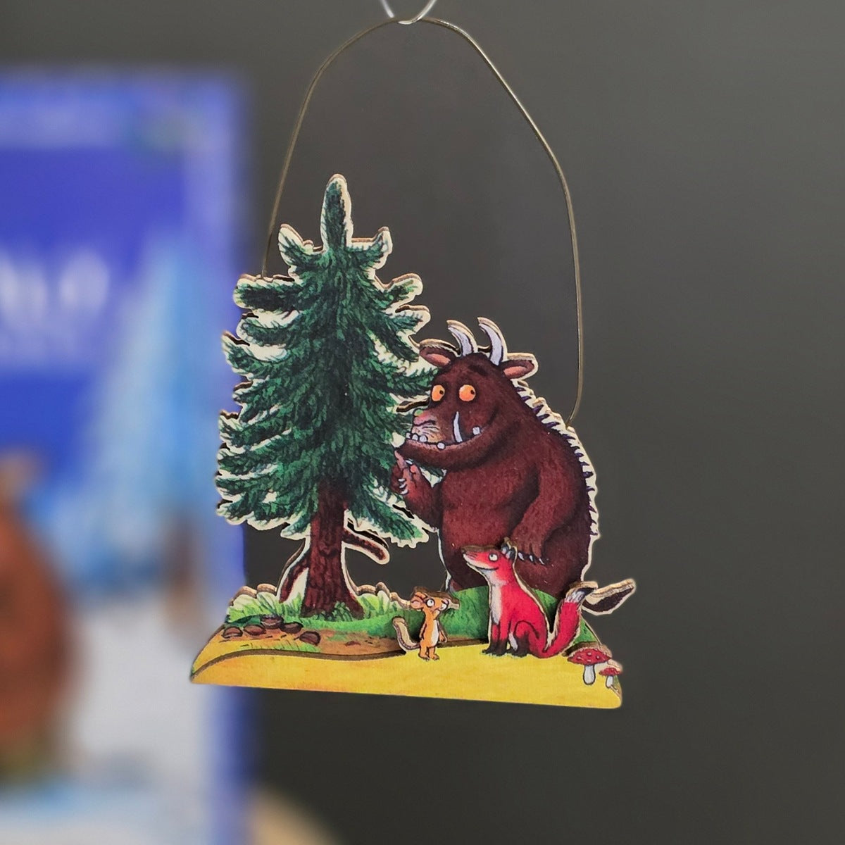 A handcrafted wooden hanging ornament featuring a whimsical illustration of the Gruffalo, a red fox, and a pine tree on a yellow forest floor, suspended by a thin metal wire against a dark grey background.