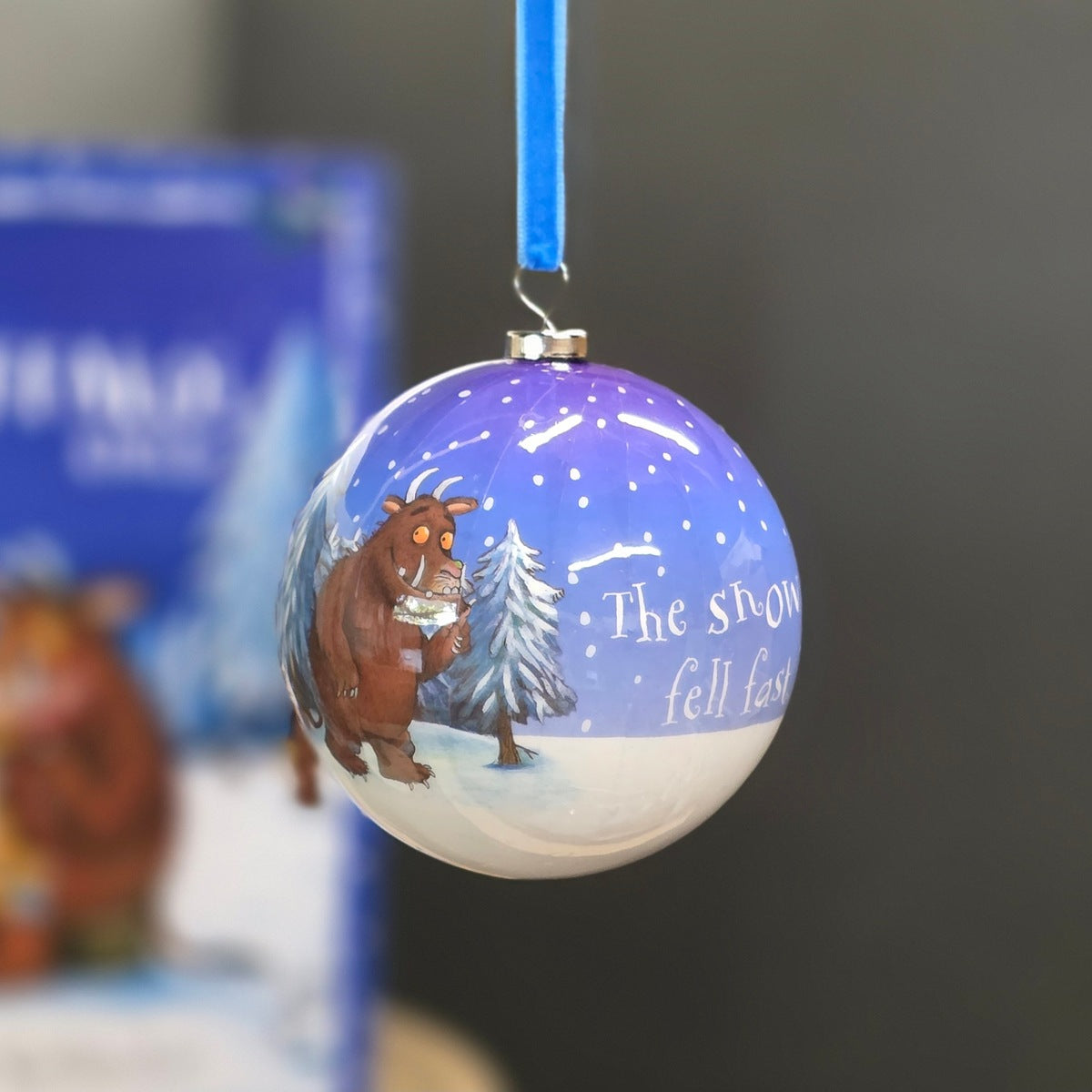 A round Christmas bauble featuring a winter scene with the Gruffalo standing beside a snowy tree under a purple sky, with the text 'The snow fell last night.' A blue hanging ribbon is attached at the top.