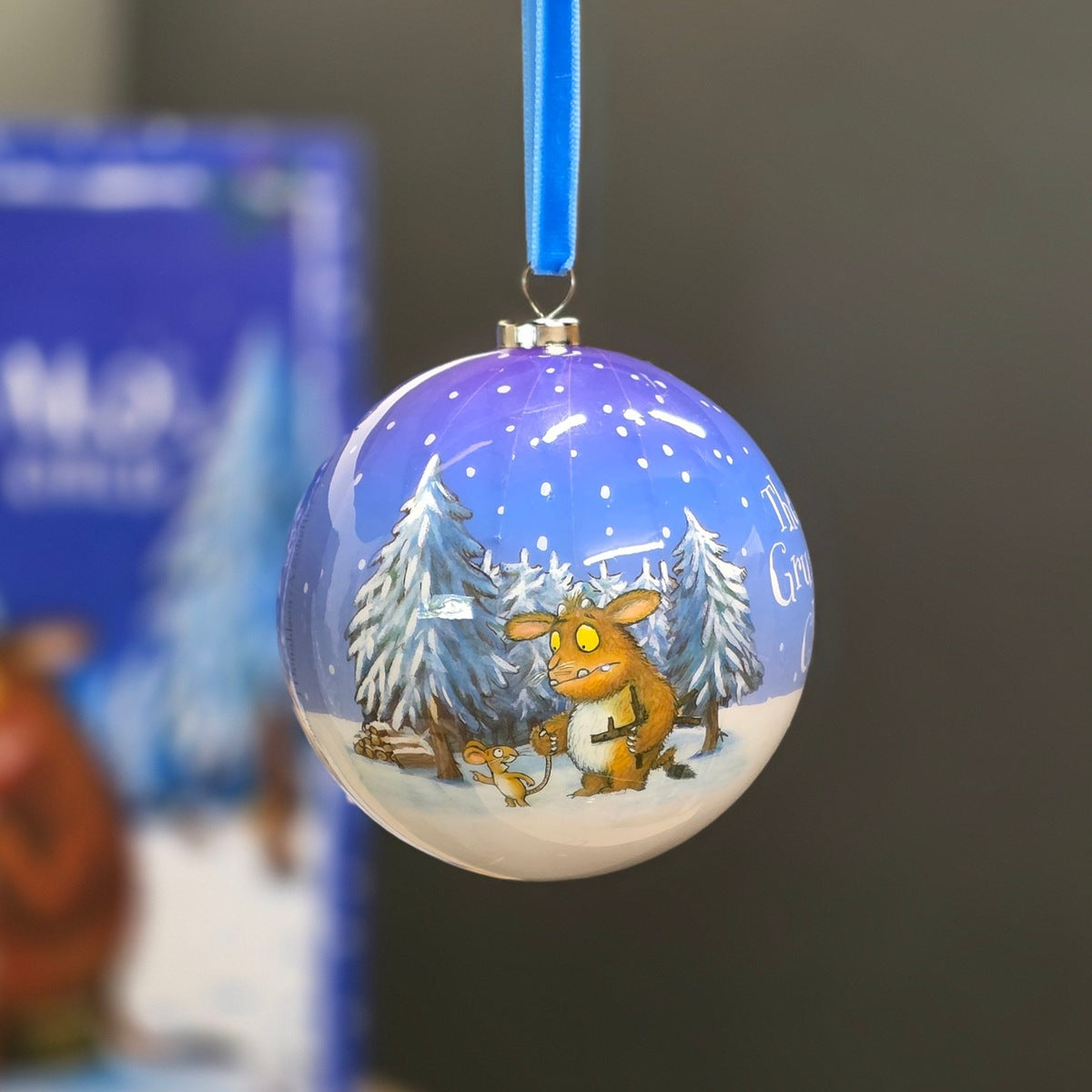 A round Christmas bauble featuring a winter forest scene with a Gruffalo and a mouse under a blue starry sky, hanging by a blue ribbon against a blurred background.