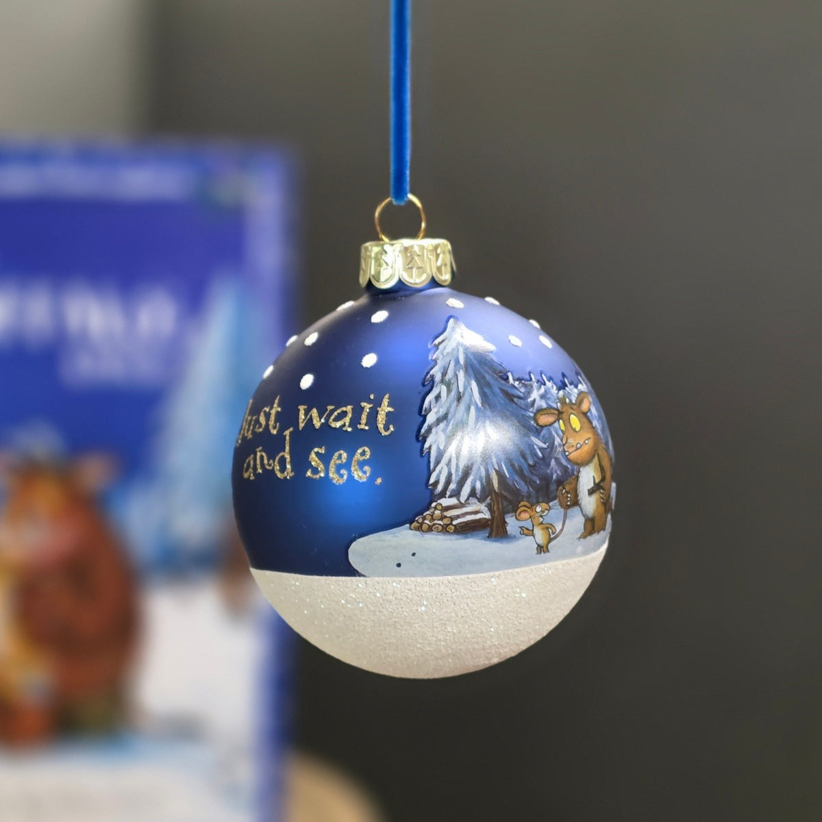 A hand-painted Christmas ornament hanging by a blue ribbon. The ornament is split horizontally, with a dark blue top half featuring white snowflakes, gold text reading 'just wait and see.' with an image of a Gruffalo and a mouse.