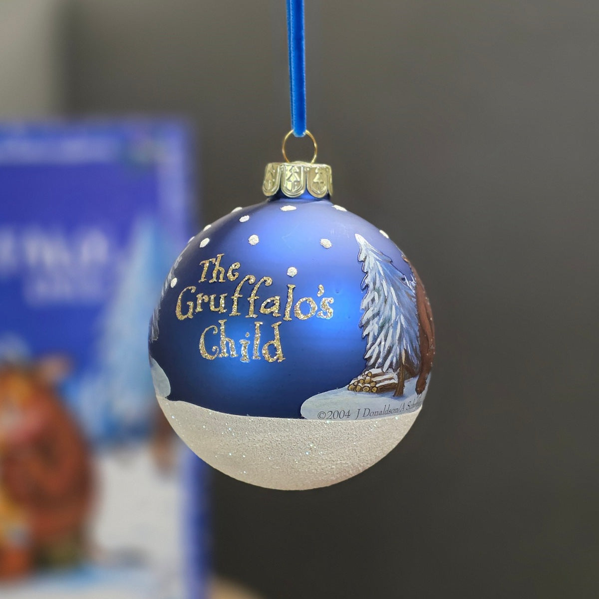 A blue and white Christmas bauble featuring the text 'The Gruffalo's Child' in gold, with a gold tree and brown Gruffalo illustration, hanging by a blue ribbon.