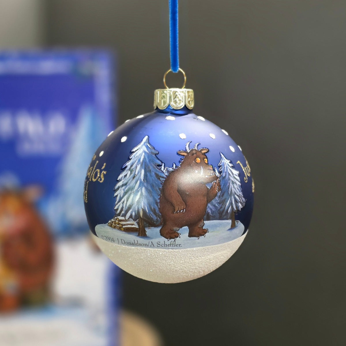 A hand-painted Christmas ornament hanging by a blue ribbon. The ornament is split horizontally, with a dark blue top half featuring white snowflakes,  with an image of a Gruffalo.