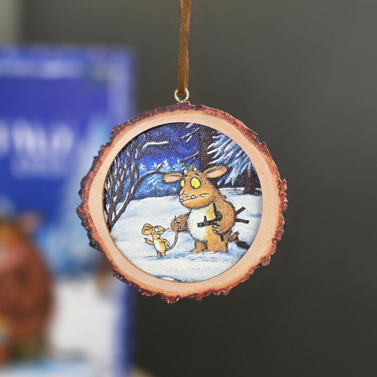 A circular wood slice ornament featuring a painted winter scene with a Gruffalo creature and a small yellow mouse in the snow, hanging by a brown ribbon against a blurred background.