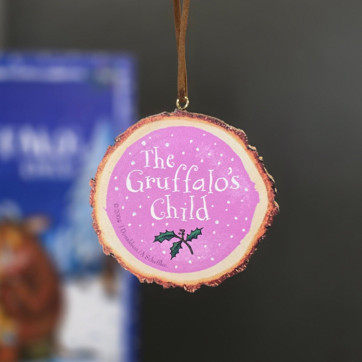 A round wooden Christmas ornament hanging from a brown ribbon, featuring a pink background with white text reading 'The Gruffalo's Child' and a small holly graphic.
